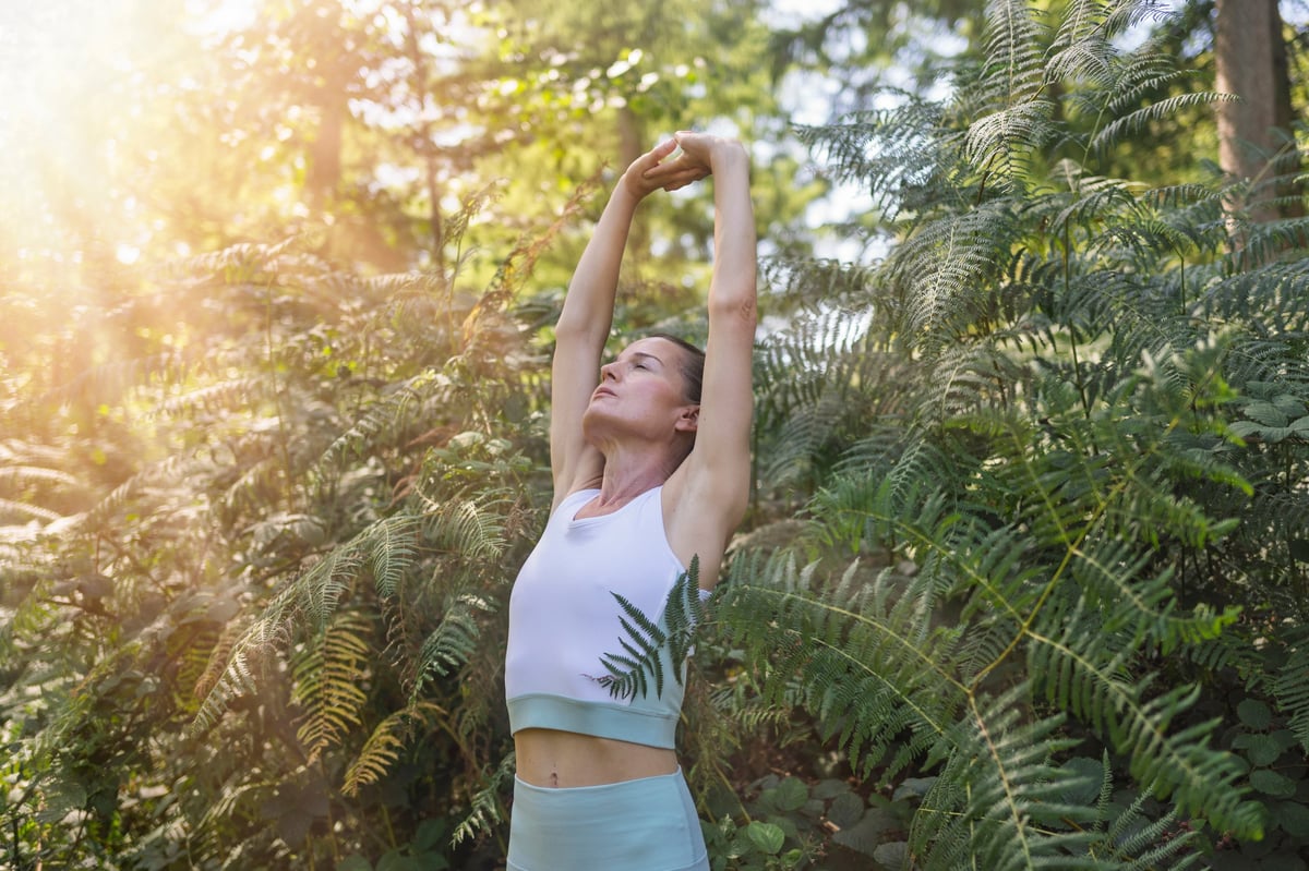 Woman practicing yoga and meditating outdoors in nature among green ferns