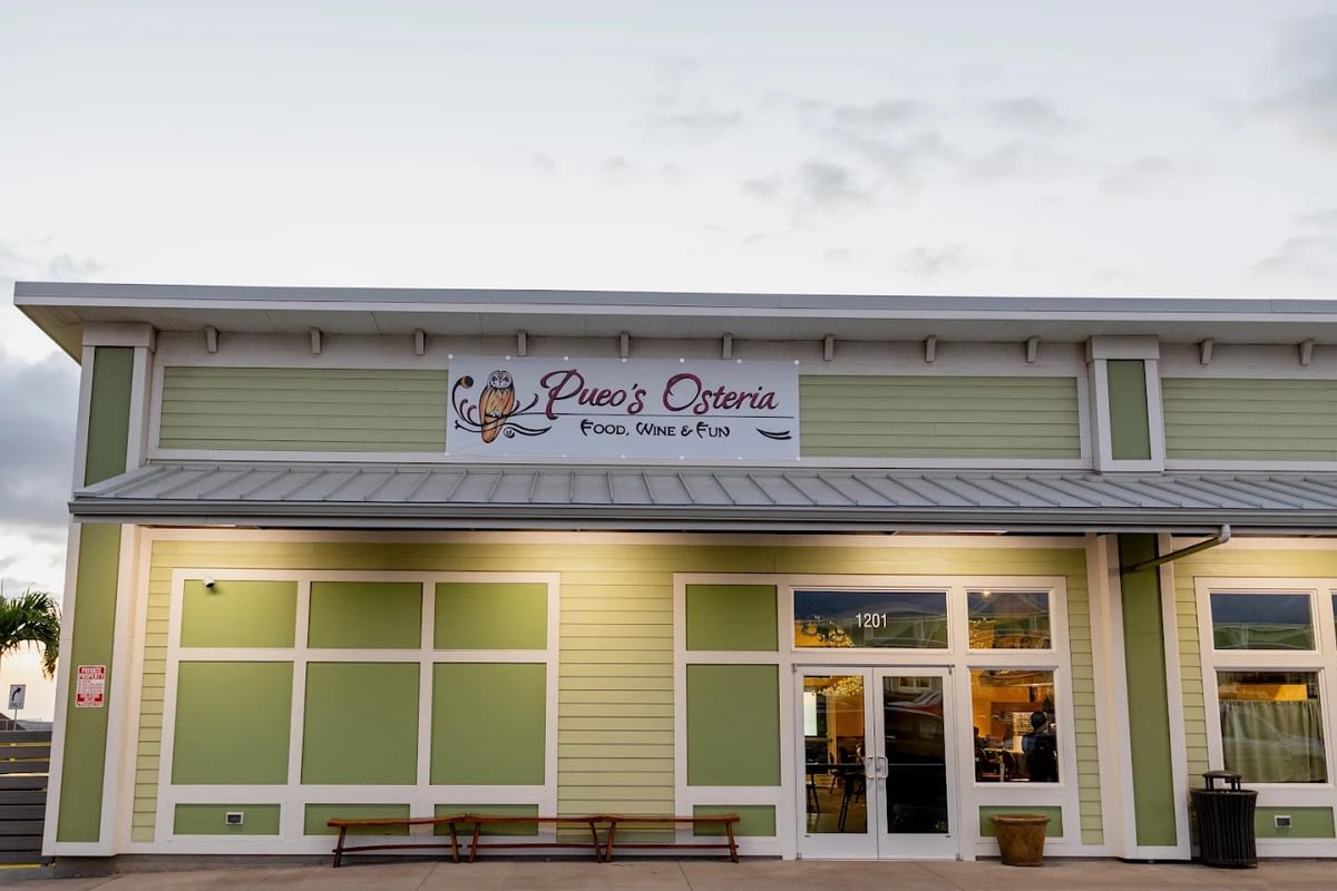 Exterior of La Pana Osteria restaurant with light green siding, storefront windows, and signage against a cloudy sky — Pueo's Osteria in Waikoloa Village