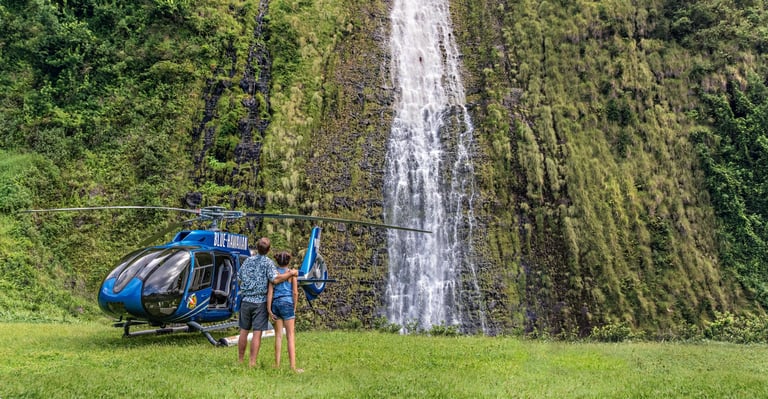 Blue helicopter parked on grass field with three people standing nearby and waterfall in background