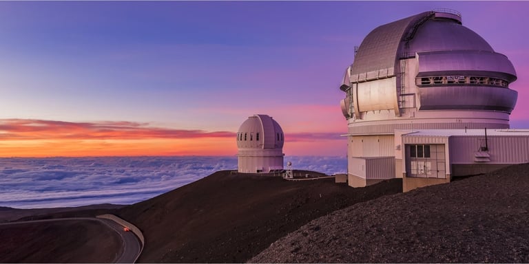 Two white domed astronomical observatories on a dark volcanic mountain ridge at sunset with colorful purple and orange sky over the ocean