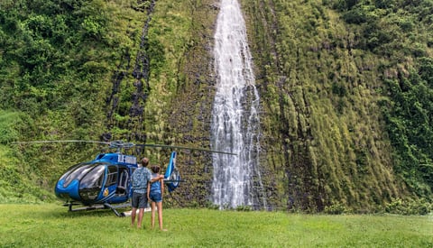 Blue helicopter on grass with large waterfall and dense green forest in background