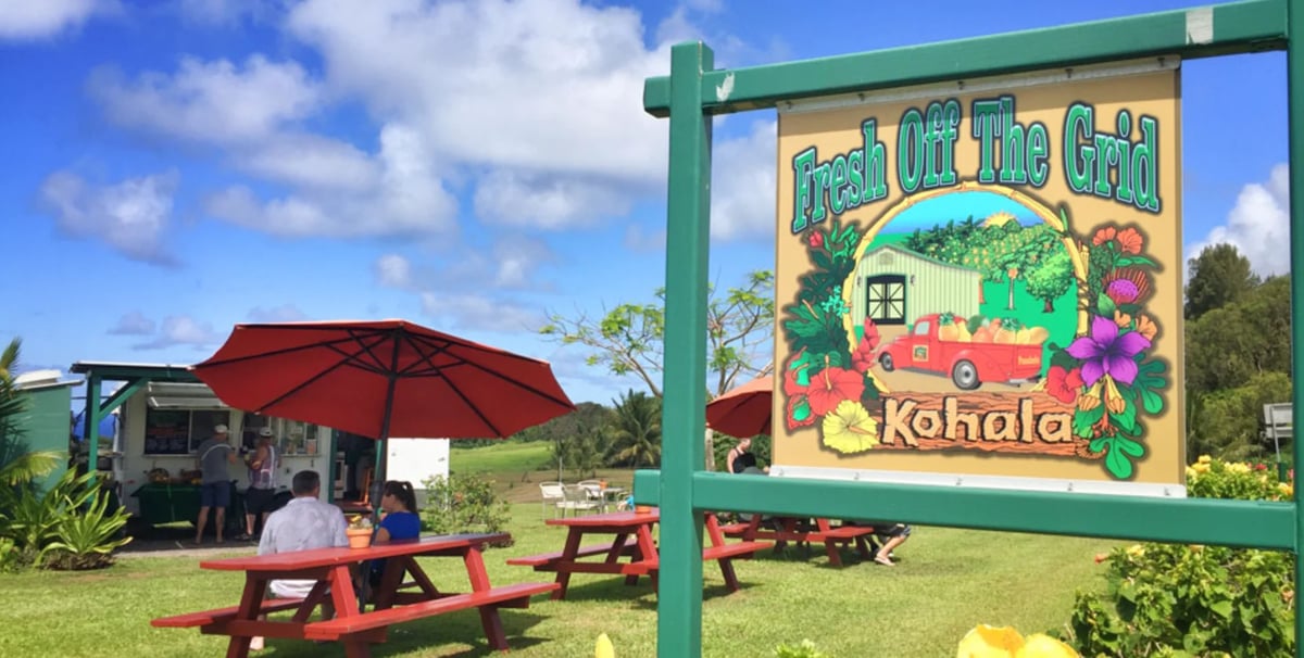 Outdoor farm stand with green sign reading Pick your own and Kahala, picnic tables, red umbrella, and gardens under blue sky — Fresh Off The Grid in Hawi North Kohala