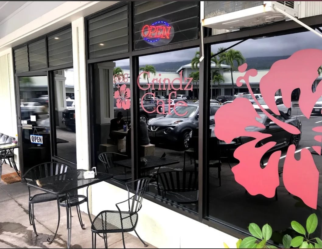 Modern storefront with black facade, large pink flower decal on window, neon signage, and black bar seating outside — 808 Grindz in Kailua-Kona