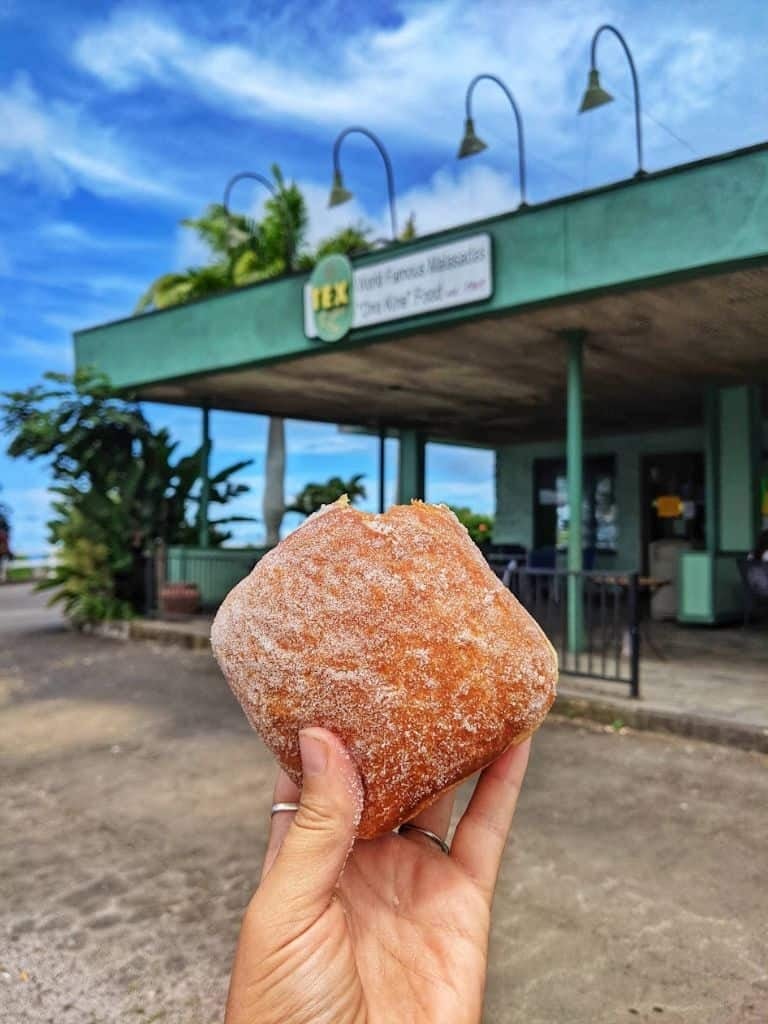 Hand holding a cinnamon sugar-coated pastry in front of a green storefront building with blue sky — Tex Drive-In in Honoka'a