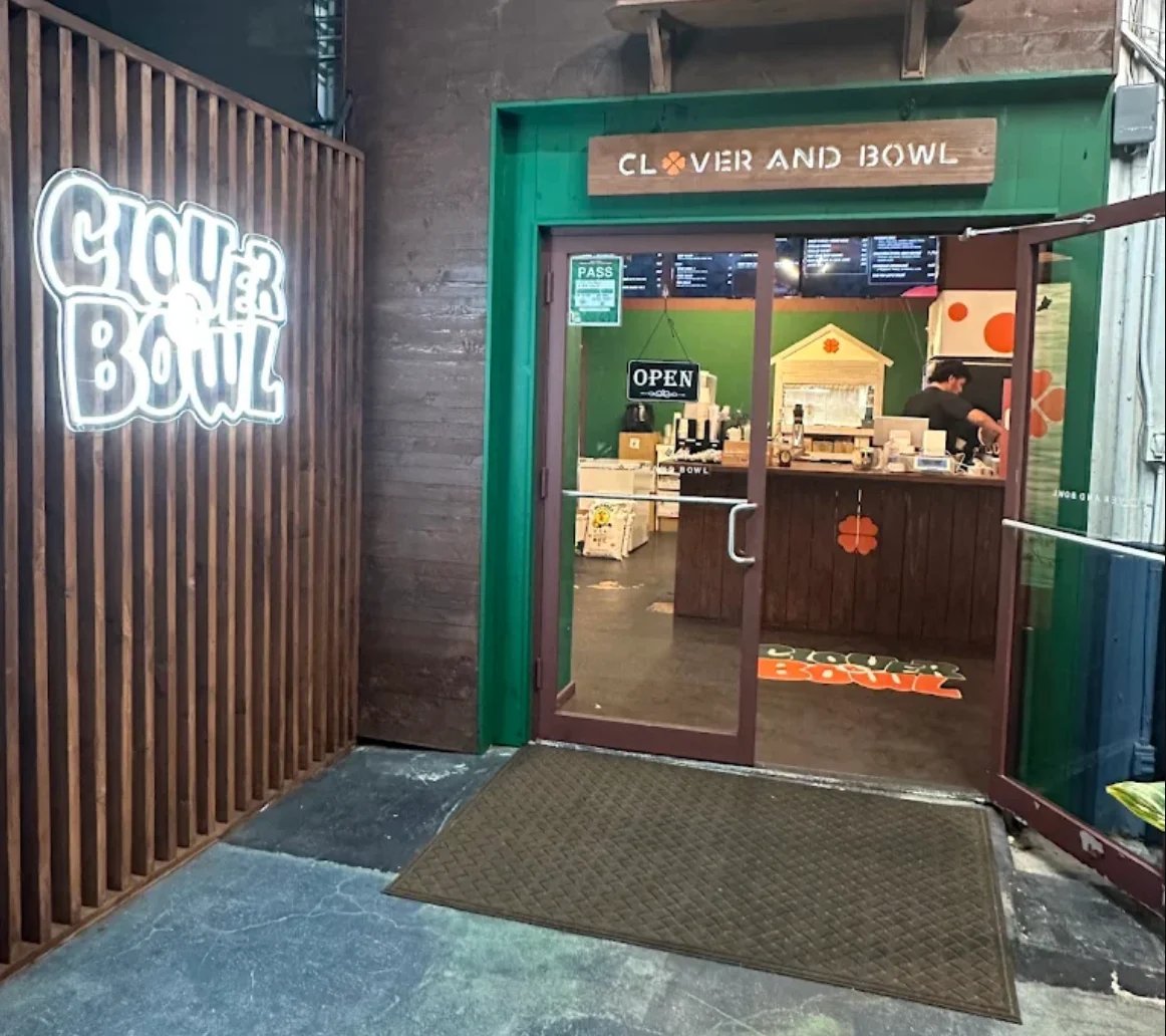 Exterior storefront with Clover and Bowl signage, neon sign, green frame entrance, and tropical plant decoration